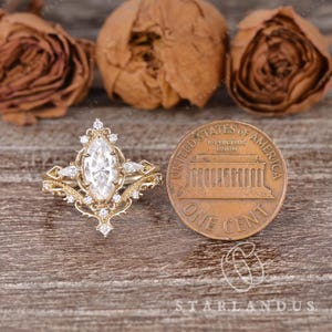 Puede incluir: Un anillo de oro con una piedra central de corte marquesa rodeada de piedras redondas y en forma de pera, junto a una moneda de un centavo de EE. UU. para escala, mostrando un trabajo de metal intrincado y un dise&ntilde;o de banda de filigrana.