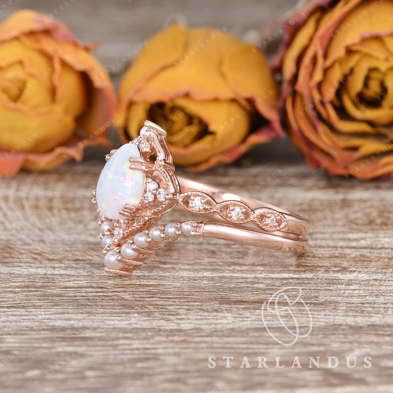 May include: A pear-shaped opal engagement ring set in rose gold with a delicate pearl and diamond band. The ring is set against a backdrop of dried roses.