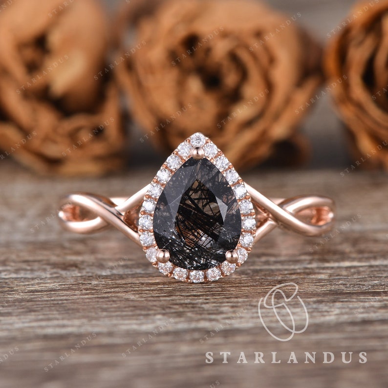 May include: A rose gold engagement ring with a pear-shaped black diamond center stone surrounded by a halo of smaller diamonds. The band is a twisted design.