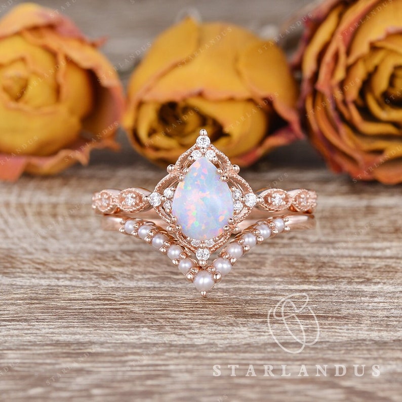 May include: A pear-shaped opal engagement ring set in rose gold with a delicate pearl and diamond band. The ring features a vintage-inspired design with intricate details.