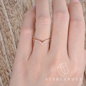 May include: A delicate rose gold ring with a V-shaped design featuring small, clear gemstones. The ring is worn on the finger of a hand.