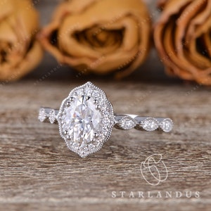 May include: A silver engagement ring with an oval-shaped center stone surrounded by a halo of smaller stones. The band is decorated with a delicate, intricate design.