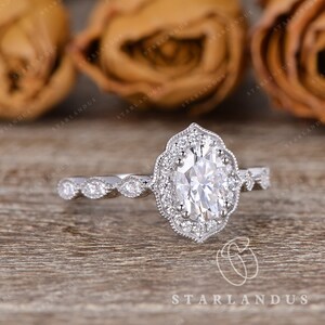 May include: A silver engagement ring with an oval-shaped center stone surrounded by a halo of smaller stones. The band is decorated with a delicate, vintage-inspired design.