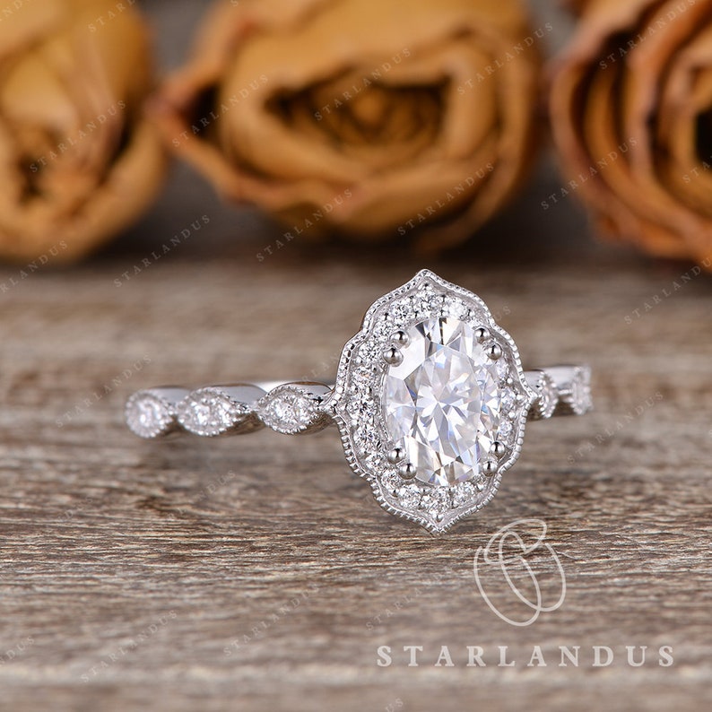 May include: A silver engagement ring with an oval-shaped center stone surrounded by a halo of smaller stones. The band is decorated with a delicate, vintage-inspired design.