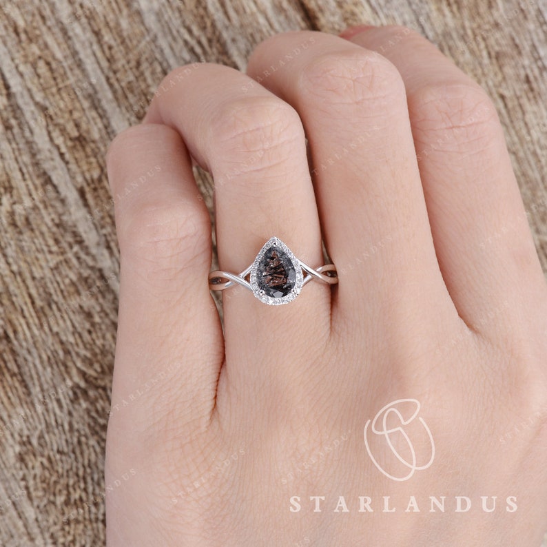 May include: A pear-shaped engagement ring with a dark grey gemstone surrounded by a halo of smaller white gemstones. The ring has a twisted band and is set in silver.