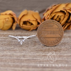 May include: A delicate, curved wedding band with a pear-shaped center stone and smaller round stones on each side. The band is set in white gold and is shown next to a US penny for size comparison.