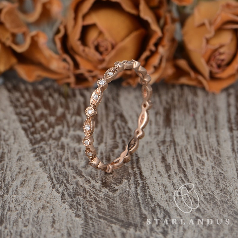 Op de afbeelding: Een delicate ros&eacute;gouden trouwring met een delicaat, ingewikkeld ontwerp met kleine ronde diamanten die in een bladvormig patroon zijn gezet.
