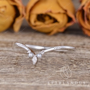 May include: A delicate silver ring with a V-shaped design, accented with small diamonds and pearls. The ring is set against a backdrop of brown roses.