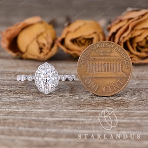 May include: A silver engagement ring with a large oval-shaped diamond set in a halo setting. The ring is surrounded by smaller diamonds and has a delicate band. The ring is shown next to a US penny for size comparison.
