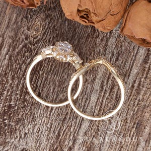 Puede incluir: Un conjunto de anillos de compromiso de oro con una piedra central de corte redondo rodeada de piedras m&aacute;s peque&ntilde;as, junto con una alianza curva a juego. Ambos anillos tienen un acabado en oro pulido y un trabajo de metal delicado.