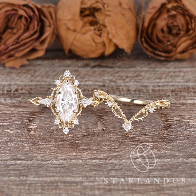 Puede incluir: Un conjunto de anillos de compromiso de oro con una piedra central de talla marquesa rodeada por un halo de peque&ntilde;os diamantes redondos y de talla marquesa, junto a una alianza en forma de V con filigrana y diamantes redondos.