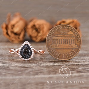 May include: A pear-shaped black diamond engagement ring with a halo of smaller diamonds set in a rose gold band. The ring is next to a US penny for size comparison.