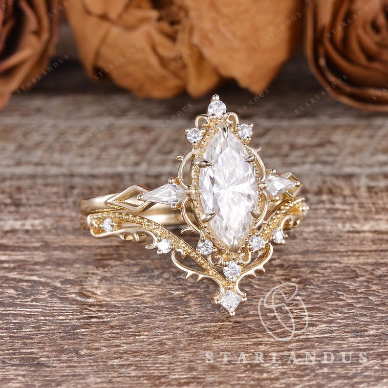 Puede incluir: Un conjunto de anillo de compromiso en oro amarillo con una piedra preciosa blanca central de corte marquesa rodeada por un halo de filigrana decorativo. El anillo incluye piedras blancas m&aacute;s peque&ntilde;as y una banda en V con detalles met&aacute;licos.
