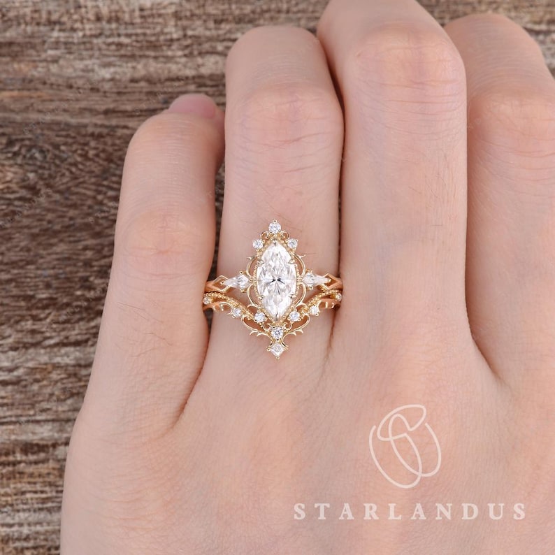Puede incluir: Un conjunto de anillos de oro con una piedra central de corte marquesa rodeada de piedras m&aacute;s peque&ntilde;as redondas y de corte marquesa en una banda decorativa de filigrana. El dise&ntilde;o incluye metalurgia intrincada y una banda en forma de V.