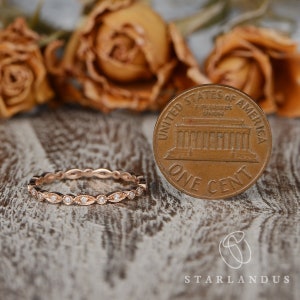 Op de afbeelding: Een delicate eeuwigheidsband van ros&eacute;goud met een rij kleine ronde diamanten die in een gegolfd ontwerp zijn gezet. De ring wordt naast een Amerikaanse cent getoond ter vergelijking van de grootte.