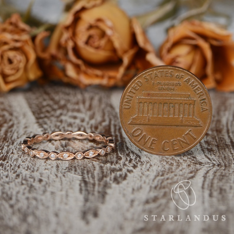 Op de afbeelding: Een delicate eeuwigheidsband van ros&eacute;goud met een rij kleine ronde diamanten die in een gegolfd ontwerp zijn gezet. De ring wordt naast een Amerikaanse cent getoond ter vergelijking van de grootte.