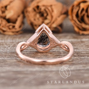 May include: A rose gold engagement ring with a pear-shaped black diamond set in a delicate, intricate design. The ring is photographed on a wooden surface with rose petals in the background.