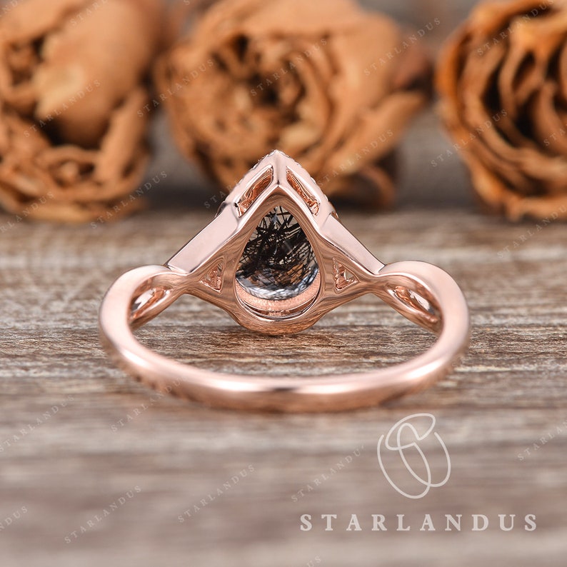 May include: A rose gold engagement ring with a pear-shaped black diamond set in a delicate, intricate design. The ring is photographed on a wooden surface with rose petals in the background.
