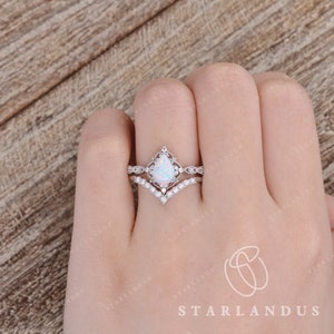 May include: A pear-shaped opal engagement ring with a delicate, ornate setting. The ring is accented with small, sparkling stones and is paired with a matching wedding band featuring a row of pearls.
