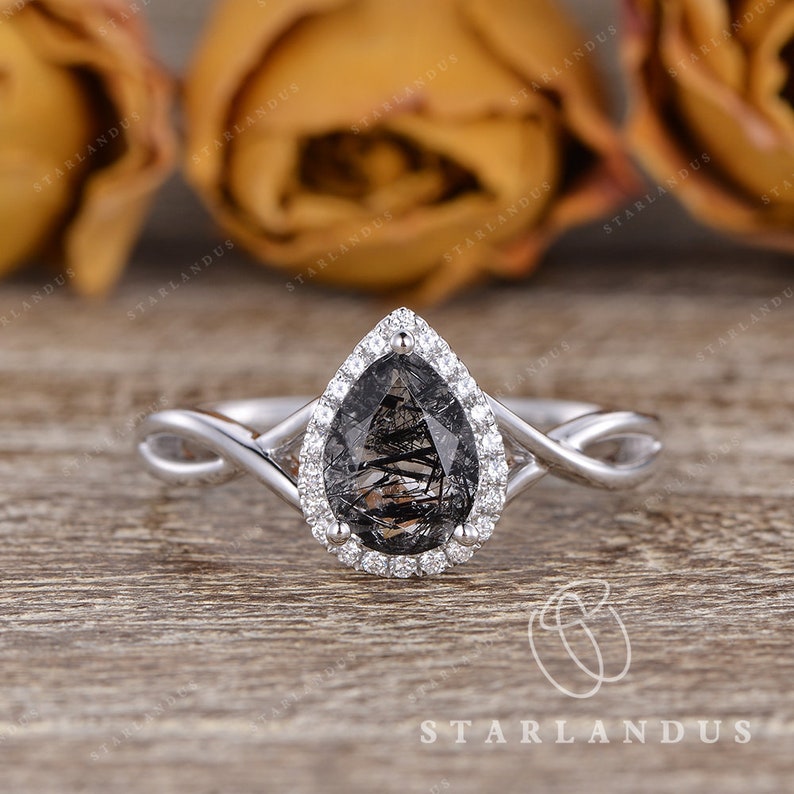 May include: A pear-shaped engagement ring with a black and silver gemstone surrounded by a halo of smaller diamonds. The ring has a twisted band.