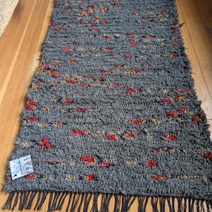 May include: A rectangular rug with a deep charcoal gray base featuring a shaggy, textured pile. The surface is interspersed with small, vibrant flecks of red, orange, and yellow yarn. The rug is finished with long, dark gray fringe at the bottom edge.