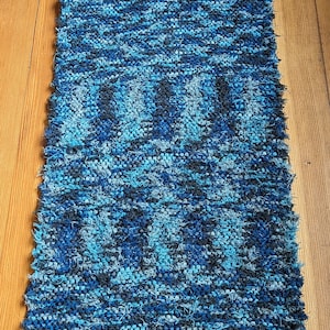 May include: A rectangular, handwoven rug with a blue and black pattern. The rug has a fringed edge and is made of a textured material. The rug is displayed on a wooden floor.