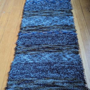 May include: A rectangular, handwoven rug with a textured surface. The rug features horizontal stripes in shades of blue, gray, and brown. The rug has a fringe on the bottom edge. The rug is approximately 60 inches long.