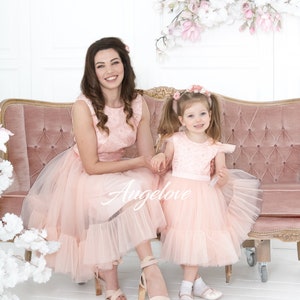 May include: A mother and daughter wearing matching pink tulle dresses with floral embroidery. The dresses have a ruffled hem and the girl's dress has a pink sash. They are sitting on a pink velvet sofa with floral decorations behind them.
