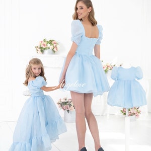 May include: Two people wearing matching light blue dresses with puffed sleeves. The dress on the left has a longer skirt with ruffles. The dress on the right has a shorter skirt. Both dresses have a square neckline and a zipper in the back.