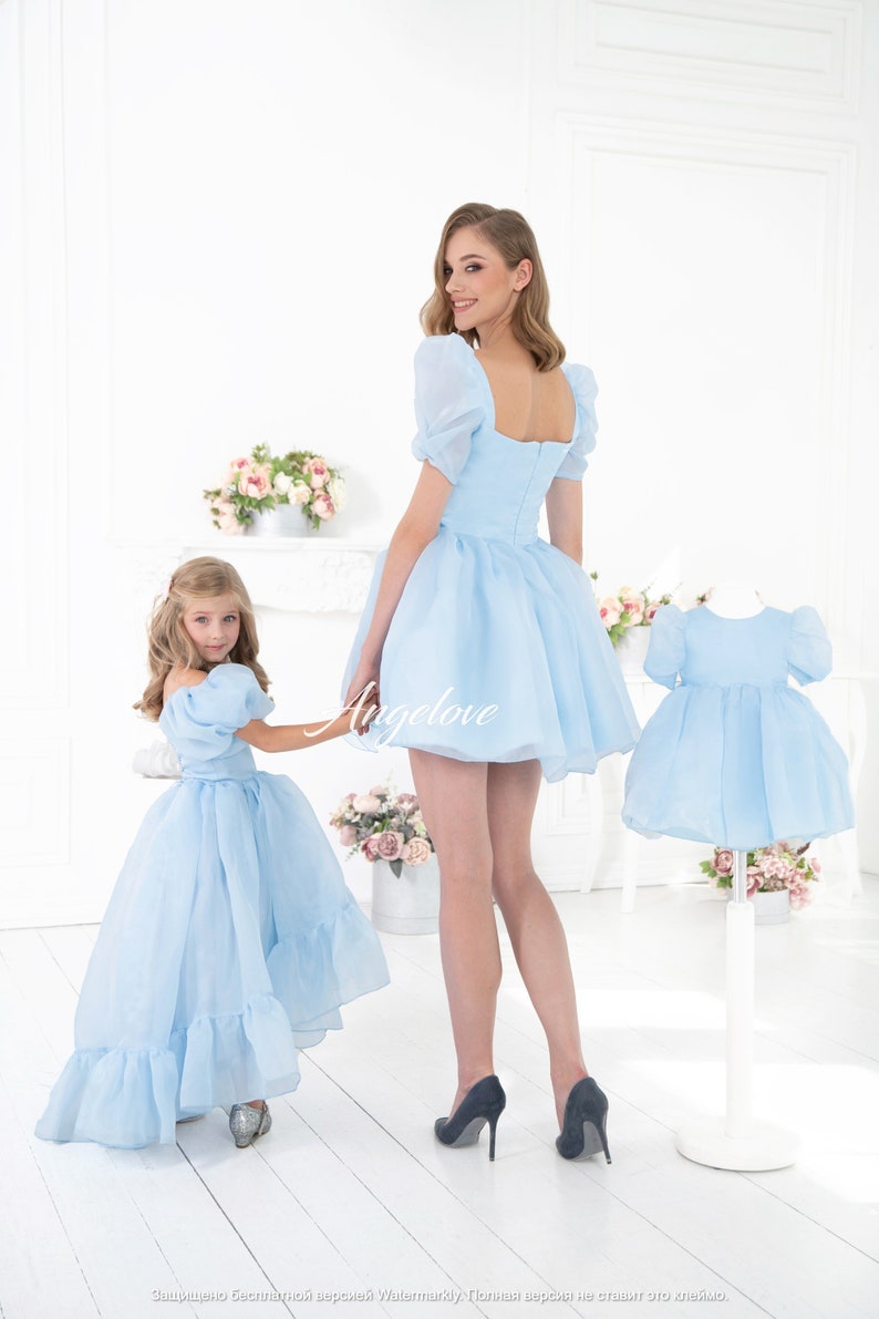 May include: Two people wearing matching light blue dresses with puffed sleeves. The dress on the left has a longer skirt with ruffles. The dress on the right has a shorter skirt. Both dresses have a square neckline and a zipper in the back.