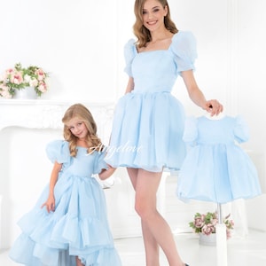 May include: Two people wearing matching light blue dresses with puffed sleeves. The dress on the left has a high-low hemline. The dress on the right is a shorter, more traditional style. Both dresses have a square neckline.