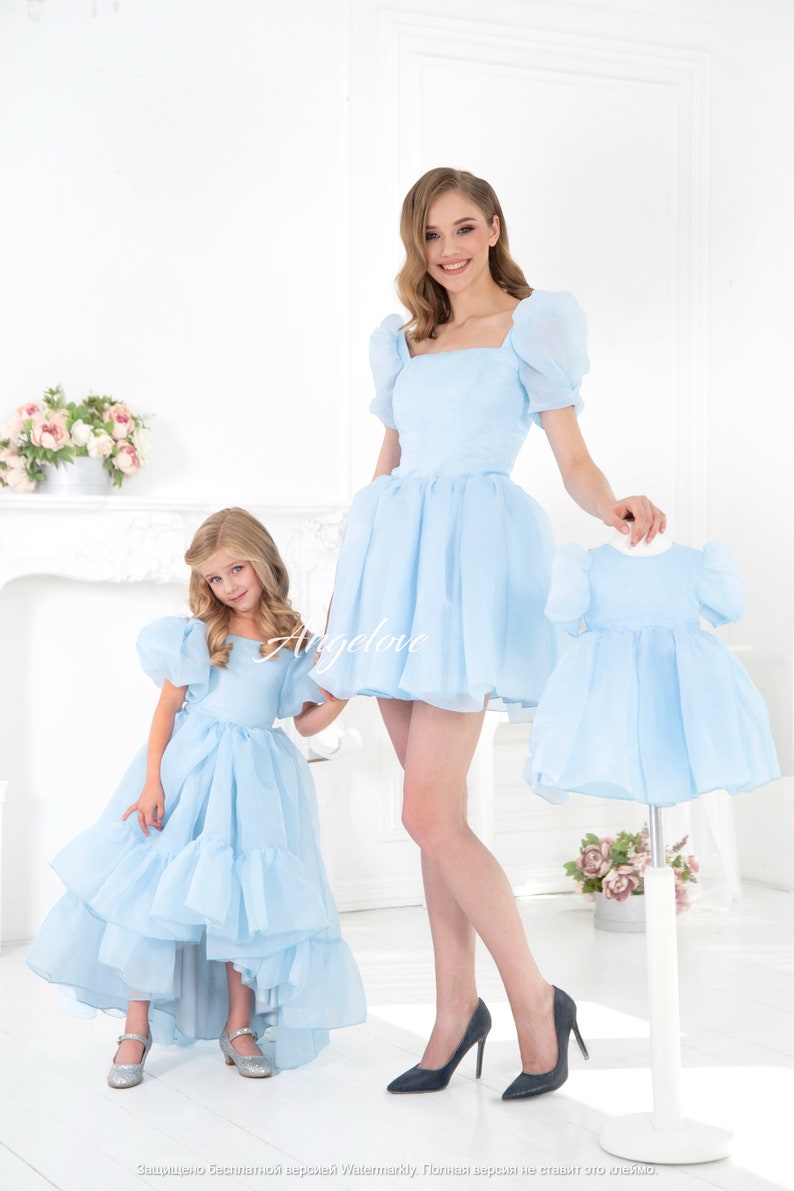 May include: Two people wearing matching light blue dresses with puffed sleeves. The dress on the left has a high-low hemline. The dress on the right is a shorter, more traditional style. Both dresses have a square neckline.