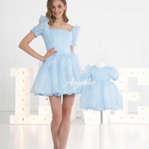 May include: Two light blue dresses, one for an adult and one for a child, with puffed sleeves and a square neckline. The dresses have a tulle skirt and are made of a soft, flowing fabric. The word "Angelove" is printed on the dress.
