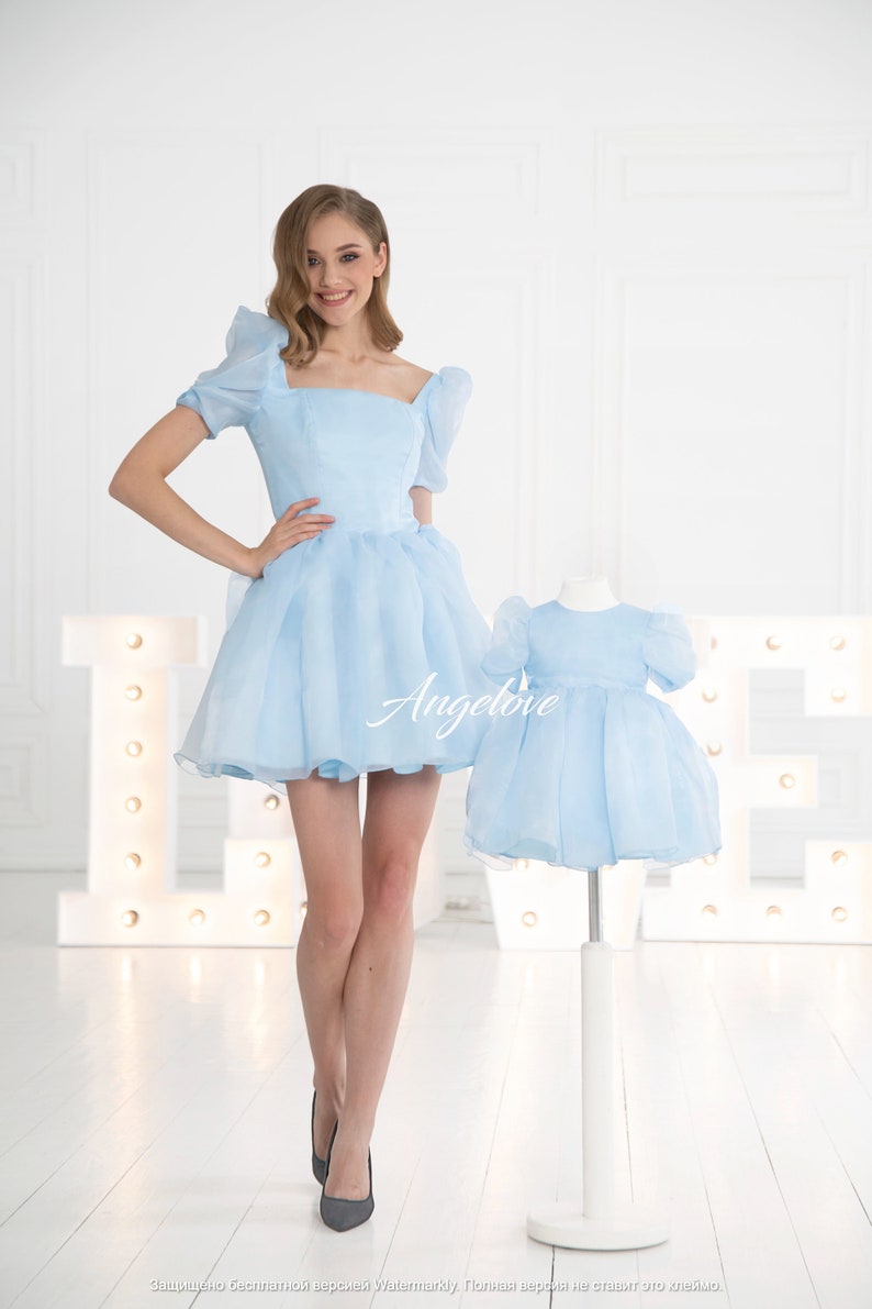 May include: Two light blue dresses, one for an adult and one for a child, with puffed sleeves and a square neckline. The dresses have a tulle skirt and are made of a soft, flowing fabric. The word "Angelove" is printed on the dress.