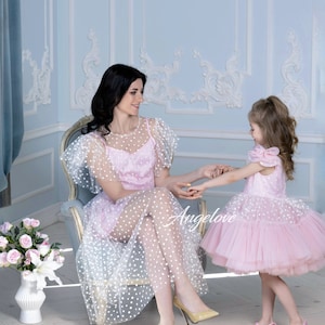 May include: A mother and daughter are pictured in a room, both wearing matching pink dresses with white polka dots. The woman is seated, and the girl is standing, holding hands. The dresses have tulle skirts. The word "Angelove" is visible.