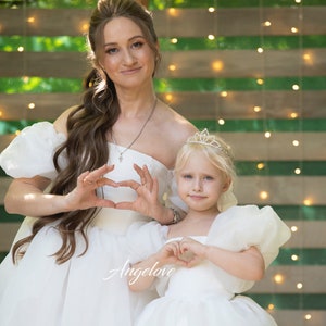 May include: Two people wearing white dresses, one with a crown, are making heart shapes with their hands. The woman is holding the girl in her arms. The background is a green and white striped wall with lights.