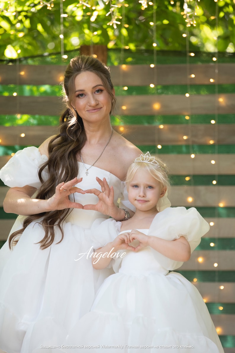 May include: Two people wearing white dresses, one with a crown, are making heart shapes with their hands. The woman is holding the girl in her arms. The background is a green and white striped wall with lights.