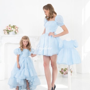 May include: Two girls wearing matching light blue dresses with puffy sleeves. The dress on the left has a high-low hemline. The dress on the right is a shorter, more traditional style. Both girls are smiling and looking at each other. The text "Angelove" is visible in the image.