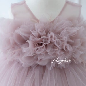 May include: A mauve tulle dress with a large, ruffled flower on the bodice. The dress has a sleeveless design and a tulle skirt. The dress is on a white mannequin.