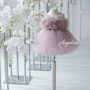 May include: A dusty rose tulle dress with a large flower detail on the bodice. The dress is on a white stand against a white background with white orchid flowers.