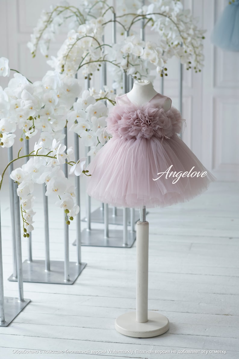 May include: A dusty rose tulle dress with a large flower detail on the bodice. The dress is on a white stand against a white background with white orchid flowers.
