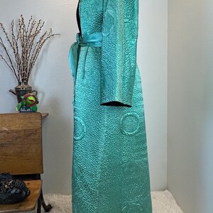K&ouml;nnte beinhalten: Ein langer, t&uuml;rkisfarbener Robe mit strukturierter Oberfl&auml;che und schwarzem Kragen. Die Robe hat einen passenden t&uuml;rkisfarbenen G&uuml;rtel und dekorative kreisf&ouml;rmige Muster. Das Kleidungsst&uuml;ck wird auf einer Schaufensterpuppe pr&auml;sentiert.
