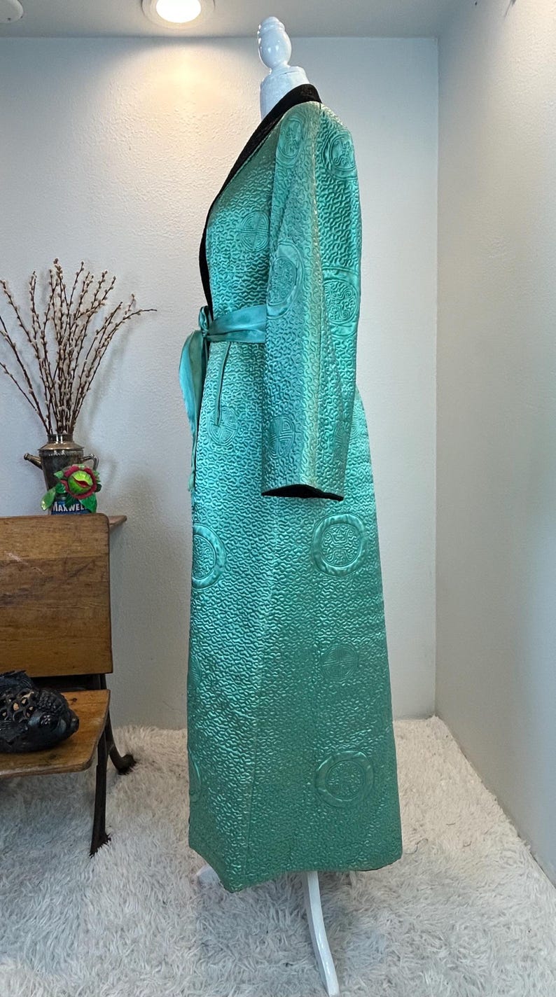 K&ouml;nnte beinhalten: Ein langer, t&uuml;rkisfarbener Robe mit strukturierter Oberfl&auml;che und schwarzem Kragen. Die Robe hat einen passenden t&uuml;rkisfarbenen G&uuml;rtel und dekorative kreisf&ouml;rmige Muster. Das Kleidungsst&uuml;ck wird auf einer Schaufensterpuppe pr&auml;sentiert.
