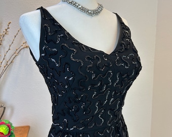 May include: A black sleeveless dress with a V-neckline and a pattern of black sequins. The dress is fitted and has a skirt that falls to the knees.