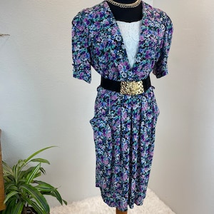 May include: A floral print dress with a V-neckline, short sleeves, and a black belt with a gold buckle. The dress is on a mannequin and has a white lace top underneath.