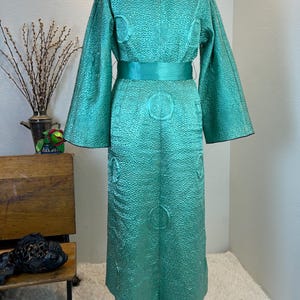 K&ouml;nnte beinhalten: Ein Vintage-Kimono-&auml;hnlicher Robe in T&uuml;rkis mit einem strukturierten Muster und einem passenden G&uuml;rtel. Die Robe hat weite &Auml;rmel und einen schwarzen Kragen. Das Design umfasst kreisf&ouml;rmige Verzierungen. Die Robe wird auf einer Schaufenquin pr&auml;sentiert.