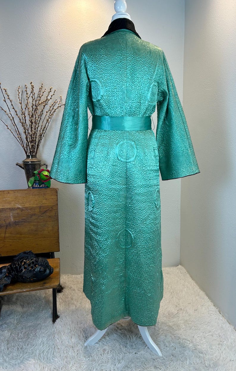 K&ouml;nnte beinhalten: Ein Vintage-Kimono-&auml;hnlicher Robe in T&uuml;rkis mit einem strukturierten Muster und einem passenden G&uuml;rtel. Die Robe hat weite &Auml;rmel und einen schwarzen Kragen. Das Design umfasst kreisf&ouml;rmige Verzierungen. Die Robe wird auf einer Schaufenquin pr&auml;sentiert.