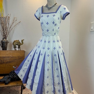 May include: A white dress with short sleeves and a square neckline, featuring a repeating purple embroidered floral pattern. The skirt is designed with alternating white and solid purple vertical pleated panels, creating a full, flared silhouette.