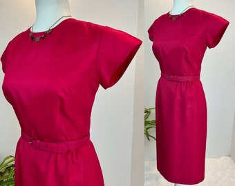 May include: A vibrant fuchsia-colored dress with short sleeves and a matching belt. The dress is displayed on a mannequin, showcasing its tailored fit. A delicate necklace adds a touch of elegance. The dress is a classic, vintage style.