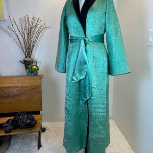 K&ouml;nnte beinhalten: Eine lange, teal-farbene Robe mit strukturiertem Muster und schwarzem Kragen wird auf einer Schaufensterpuppe pr&auml;sentiert. Die Robe hat einen passenden teal-farbenen Sch&auml;rpe, die in der Taille gebunden ist. Die &Auml;rmel sind lang und weit. Es ist ein Vintage-Kleidungsst&uuml;ck.